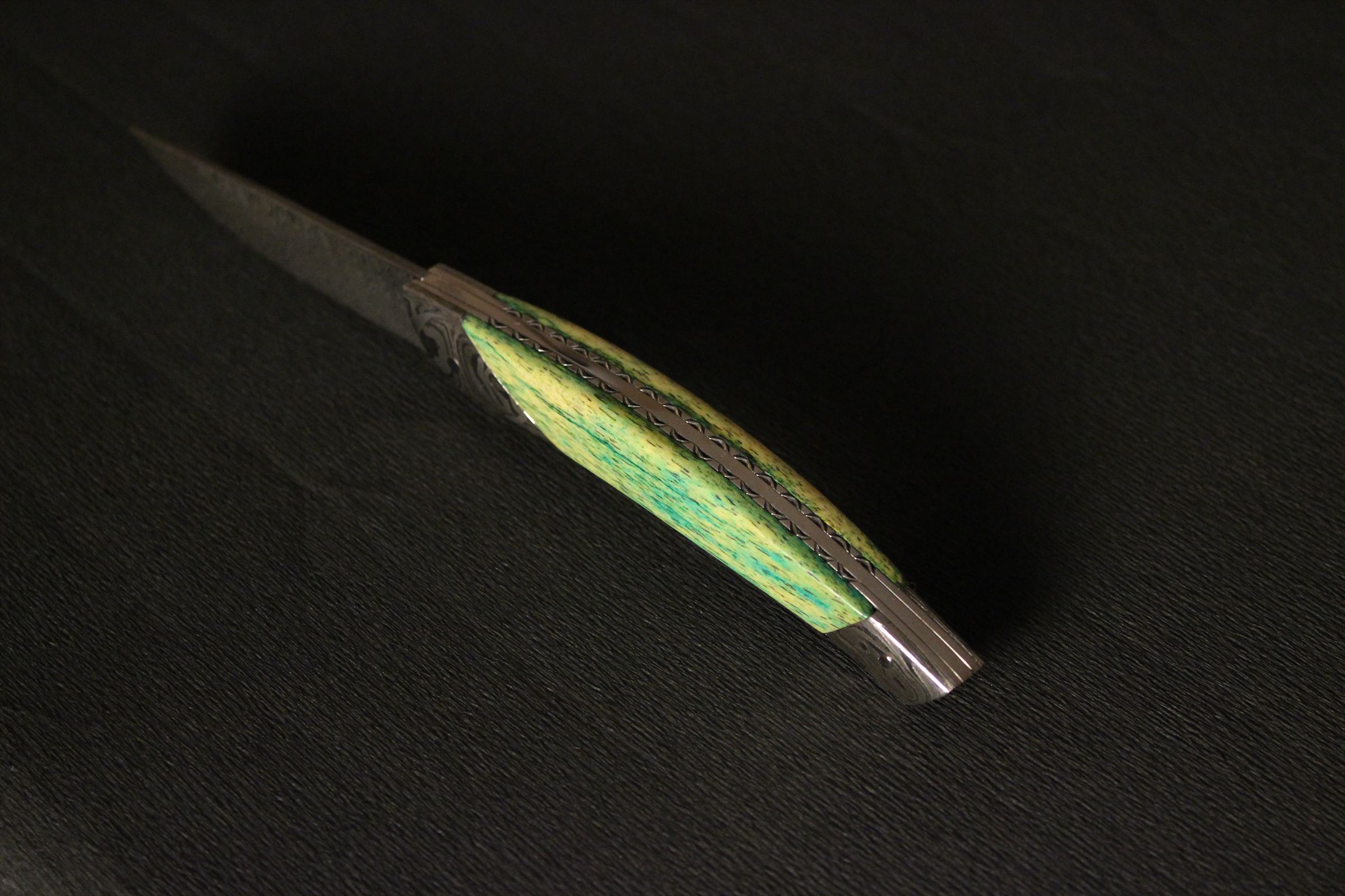 Couteau à lame damassée et manche stabilisé vert et jaune, posé sur fond noir