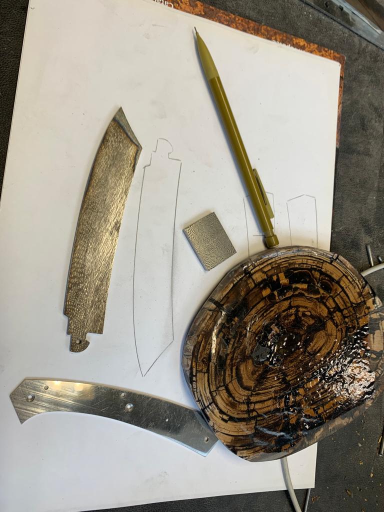 Contour d'une lame tracé au crayon sur une feuille blanche, entouré de pièces en cours de fabrication : une lame, une platine et une plaquette de manche